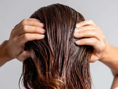 Woman applying hair mask treatment to wet brown hair. Hands massaging nourishing conditioner for deep hair repair and hydration. Professional haircare routine for damaged hair restoration.